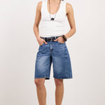 A model stands against a plain white studio background wearing blue denim shorts, a white sleeveless top, black sandals, and layered necklaces.