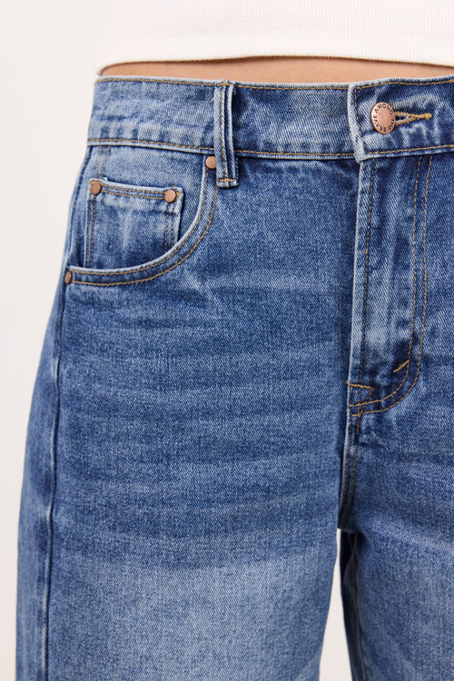 Close-up of a pair of medium-wash blue denim jeans showing the front waistband, belt loops, right front pocket, and rivets, worn with a white top against a light neutral background.
col_pattern
