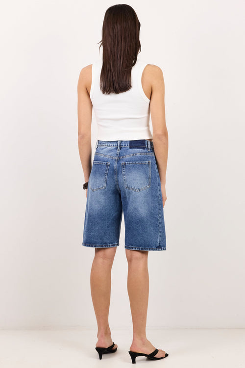 A woman viewed from behind wearing blue denim shorts, a white sleeveless top, and black heeled sandals against a plain white studio background.