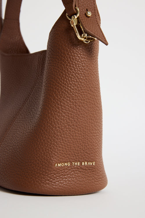A close-up of a brown pebbled leather handbag with gold hardware and the text 'AMONG THE BRAVE' printed in gold near the bottom, set against a plain light background.