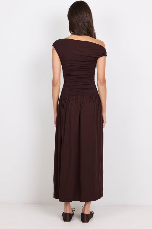 A woman viewed from the back wearing a dark brown off-the-shoulder maxi dress with a ruched bodice and flowing skirt, standing against a plain white studio background.