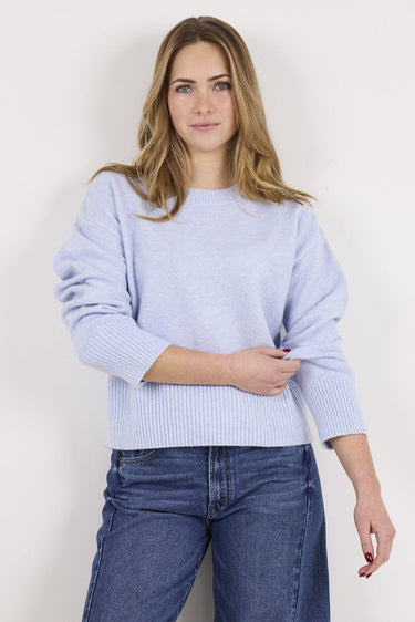 A woman modeling a light periwinkle blue knit sweater with wide three-quarter sleeves and a ribbed hem, paired with dark blue wide-leg jeans, against a plain off-white background.