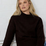 Woman wearing a black sweater and blue jeans sitting against a white brick wall.