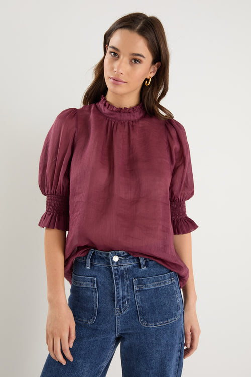 Woman wearing a burgundy blouse with puffed sleeves and blue jeans on a plain background