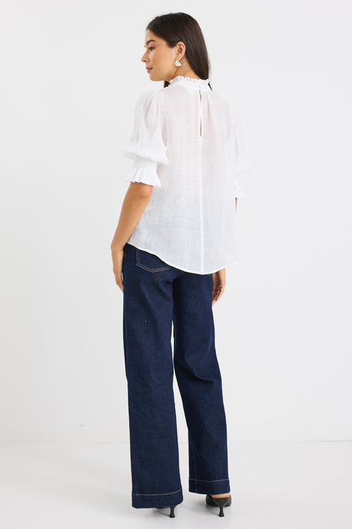 woman wearing a relaxed ivory short sleeve top with frilled neckline and sleeves, paired with indigo jeans.