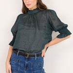 A woman modeling a dark gray sheer puff-sleeve blouse with a high gathered neckline, paired with blue wide-leg jeans and a black belt, against a plain off-white background.