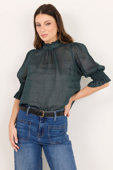 A woman modeling a dark gray sheer puff-sleeve blouse with a high gathered neckline, paired with blue wide-leg jeans and a black belt, against a plain off-white background.