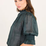A woman wearing a dark green sheer puff-sleeve blouse with a smocked elbow and ruffled hem, paired with blue jeans, photographed against a plain light background.