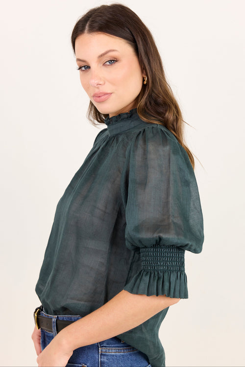 A woman wearing a dark green sheer puff-sleeve blouse with a smocked elbow and ruffled hem, paired with blue jeans, photographed against a plain light background.