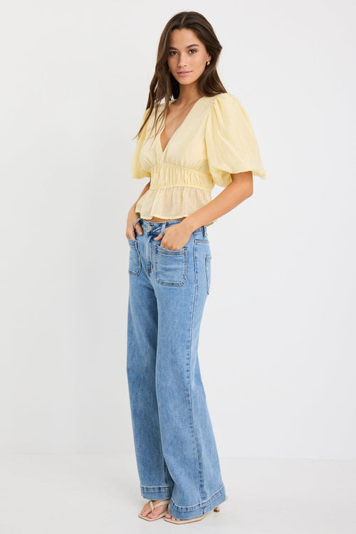 Woman wearing a butter yellow puff sleeve V-neck top with elastic shirring at the waist and a peplum hem, styled with light blue jeans.