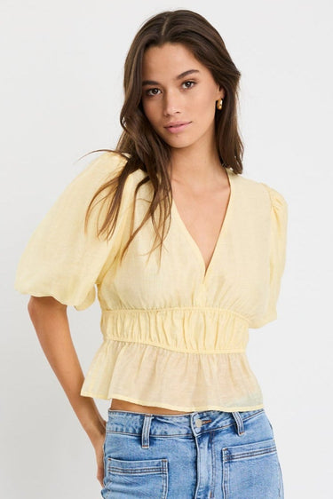 Woman wearing a butter yellow puff sleeve V-neck top with elastic shirring at the waist and a peplum hem, styled with light blue jeans.