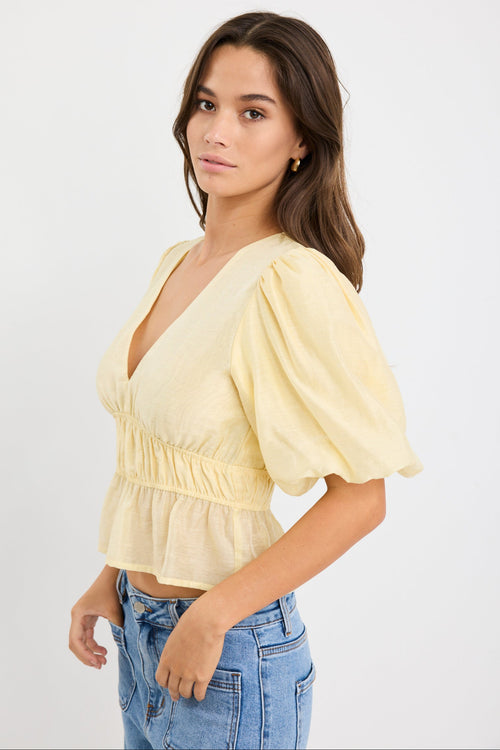Woman wearing a butter yellow puff sleeve V-neck top with elastic shirring at the waist and a peplum hem, styled with light blue jeans.