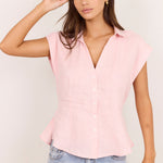 A woman modeling a light pink short-sleeve button-up blouse with a V-neck and peplum hem, paired with light-wash blue jeans against a plain off-white background.