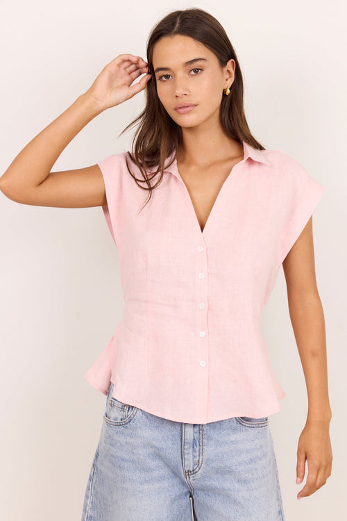 A woman modeling a light pink short-sleeve button-up blouse with a V-neck and peplum hem, paired with light-wash blue jeans against a plain off-white background.
