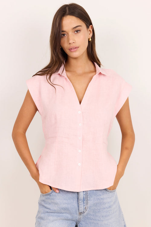 A woman modeling a light pink sleeveless button-up shirt with a V-neck and relaxed fit, paired with light-wash blue jeans against a plain off-white studio background.