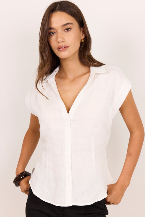 A woman modeling a white short-sleeve button-up shirt with a V-neck and collar, paired with black pants, against a plain light background.