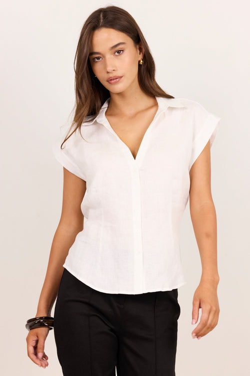 A woman modeling a white short-sleeve button-up blouse with a relaxed, slightly draped fit, paired with black trousers, against a plain light background.