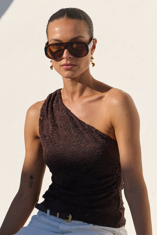 A woman wearing a dark brown textured one-shoulder top, white trousers, and sunglasses sits on a white architectural ledge in bright sunlight.