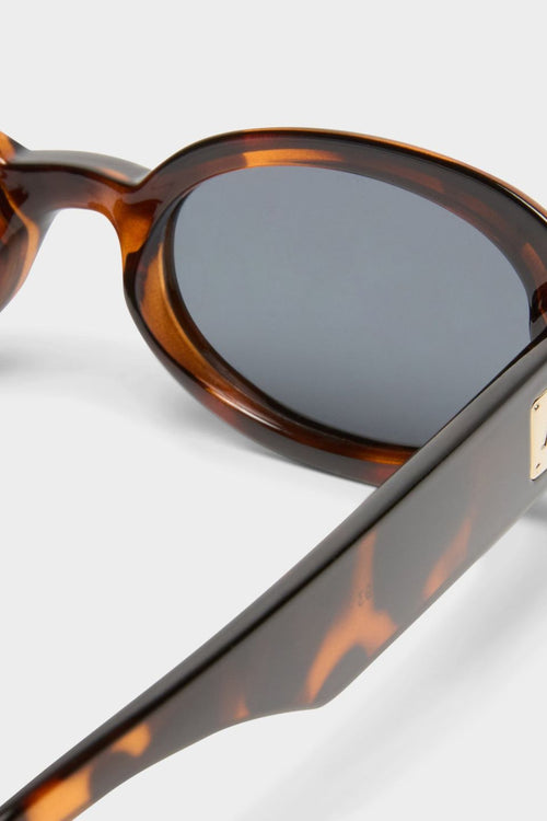 Front-facing view of oval sunglasses in a glossy dark tortoiseshell frame with smoke-tinted polarised lenses. The compact, rounded silhouette nods to 90s retro styling, offering a bold yet wearable look.