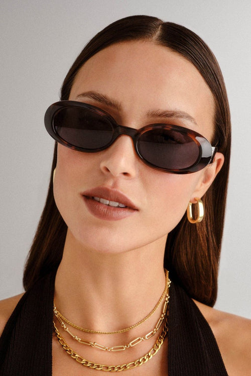 Front-facing view of oval sunglasses in a glossy dark tortoiseshell frame with smoke-tinted polarised lenses. The compact, rounded silhouette nods to 90s retro styling, offering a bold yet wearable look.