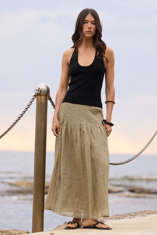 A woman stands on a concrete pier by the ocean wearing a black tank top and a long, loose beige linen skirt. The background shows the calm sea and a soft sky.