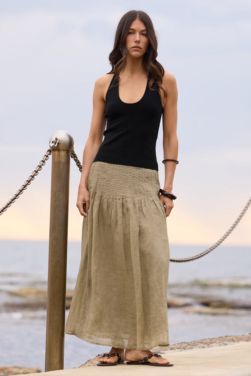 A woman stands on a concrete pier by the ocean wearing a black tank top and a long, loose khaki linen skirt. The background shows the calm sea and a soft sky.