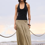 A woman stands on a concrete pier by the ocean, wearing a black sleeveless tank top and a long, loose beige maxi skirt with black sandals.