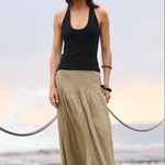 A woman stands on a concrete pier by the ocean, wearing a black sleeveless tank top and a long khaki maxi skirt with black sandals.