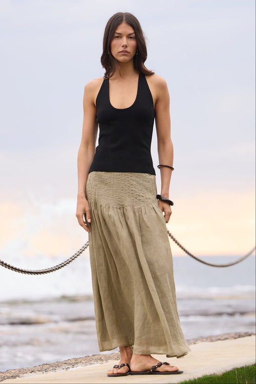 A woman stands on a concrete pier by the ocean, wearing a black sleeveless tank top and a long khaki maxi skirt with black sandals.