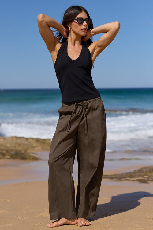 A woman stands on a sandy beach with the ocean and blue sky behind her, wearing a black sleeveless top and loose dark brown wide-leg pants.