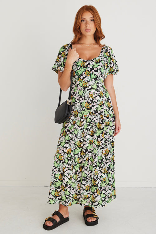 model wearing green and black floral dress and black sandals holding black handbag