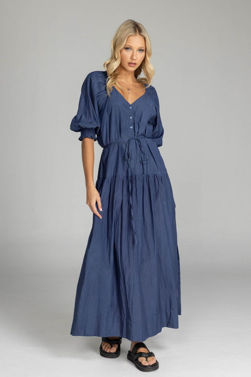 Woman wears a navy maxi dress with puff sleeves, a V-neckline, button front, waist tie, and side splits. The dress flows softly to the floor with a relaxed, feminine silhouette.