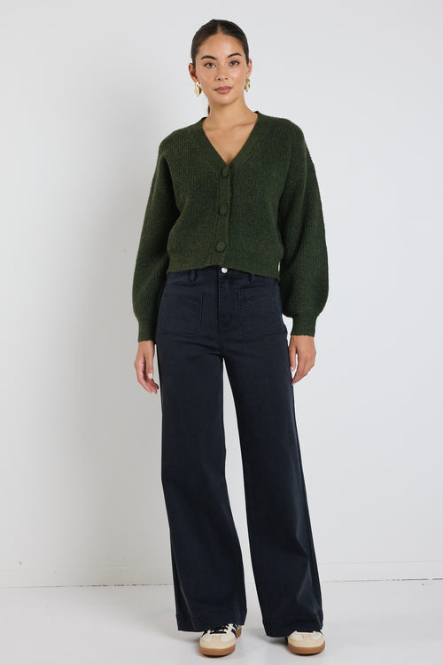 A woman wearing a dark khaki knit cardigan with oversized buttons, styled with black jeans and gold statement earrings.