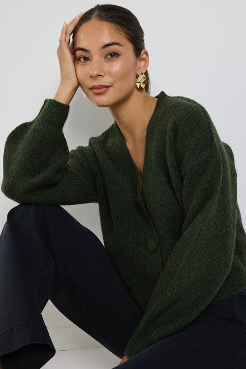 A woman wearing a dark khaki knit cardigan with oversized buttons, styled with black jeans and gold statement earrings.