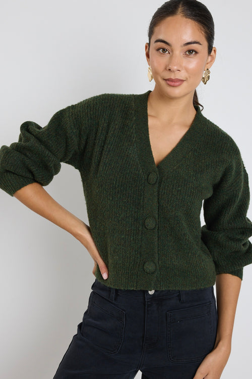 A woman wearing a dark khaki knit cardigan with oversized buttons, styled with black jeans and gold statement earrings.