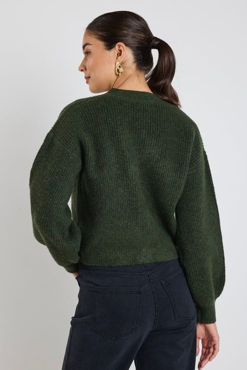 A woman wearing a dark khaki knit cardigan with oversized buttons, styled with black jeans and gold statement earrings.