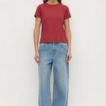 A woman stands against a plain white studio background wearing a short-sleeve crew-neck t-shirt in a muted brick red color, paired with light-wash wide-leg blue jeans and black shoes.