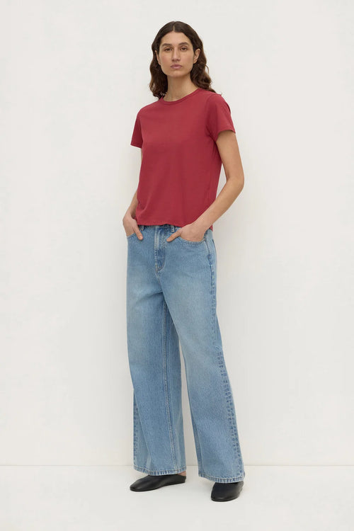 A woman modeling a short-sleeve crewneck t-shirt in dusty red and wide-leg light blue jeans, standing against a plain white studio background.