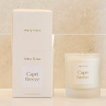 A Capri Breeze scented candle in a frosted glass jar sits on a white bathroom vanity next to its white packaging box and a dark amber glass vase with dried branches, set against a light beige tiled wall.