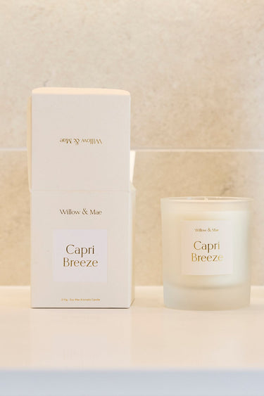 A Capri Breeze scented candle in a frosted glass jar sits on a white bathroom vanity next to its white packaging box and a dark amber glass vase with dried branches, set against a light beige tiled wall.
