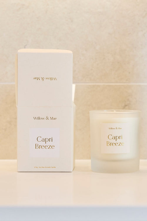 A Capri Breeze scented candle in a frosted glass jar sits on a white bathroom vanity next to its white packaging box and a dark amber glass vase with dried branches, set against a light beige tiled wall.