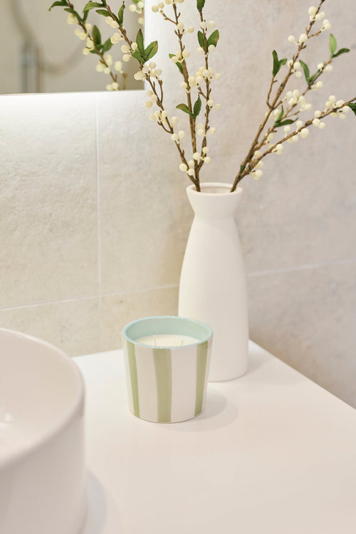 A bathroom vanity scene with a white ceramic vessel holding delicate flowering branches, next to a small striped soap dish on a white countertop.