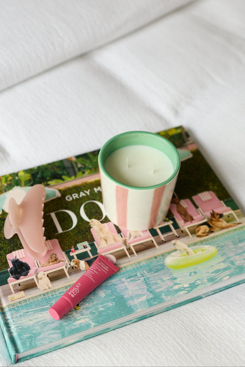 A styled product shot featuring a gray and pink striped candle with a mint green rim, resting on top of a colorful magazine on a white textured fabric surface.