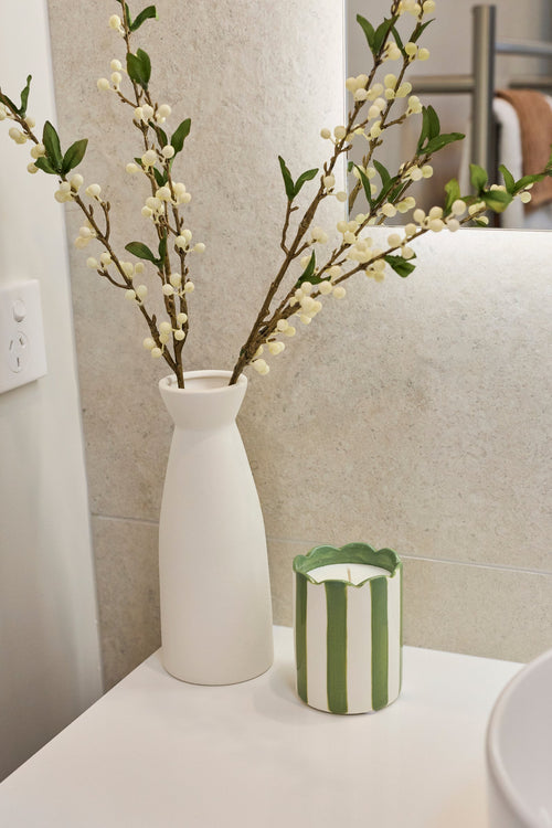 A small green and white striped candle sits on a white bathroom counter next to a white ceramic vase holding white blossoms, with a white vessel sink in the foreground.