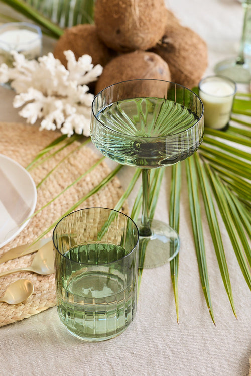A styled dining table setting featuring light green tinted glassware, woven straw placemats, white plates, and tropical green palm leaves, with coconuts and white coral as centerpiece accents.