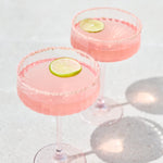Two pink margarita cocktails in stemmed coupe glasses with salted rims and lime slices