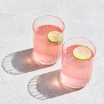 Two clear glasses filled with pink beverage and each topped with a lime wheel, placed on a light stone surface with strong sunlight and shadows.