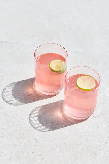 Two clear glasses filled with pink beverage and each topped with a lime wheel, placed on a light stone surface with strong sunlight and shadows.