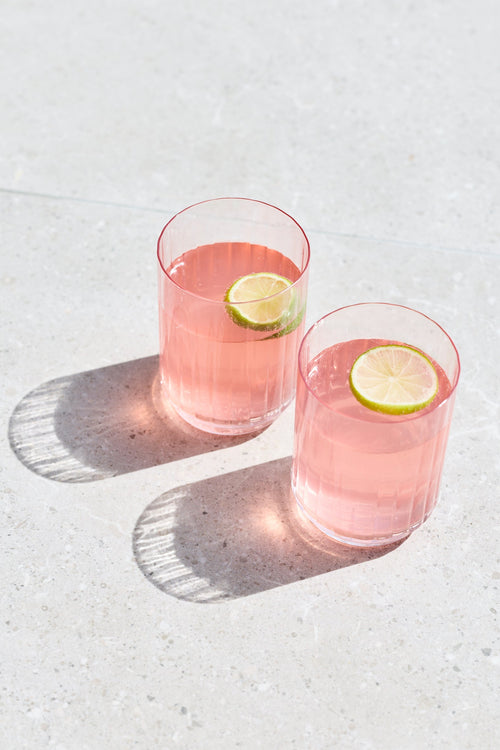Two clear glasses filled with pink beverage and each topped with a lime wheel, placed on a light stone surface with strong sunlight and shadows.