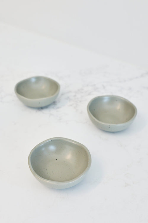 Three small shallow ceramic bowls in a muted sage green color, arranged on a white marble countertop.